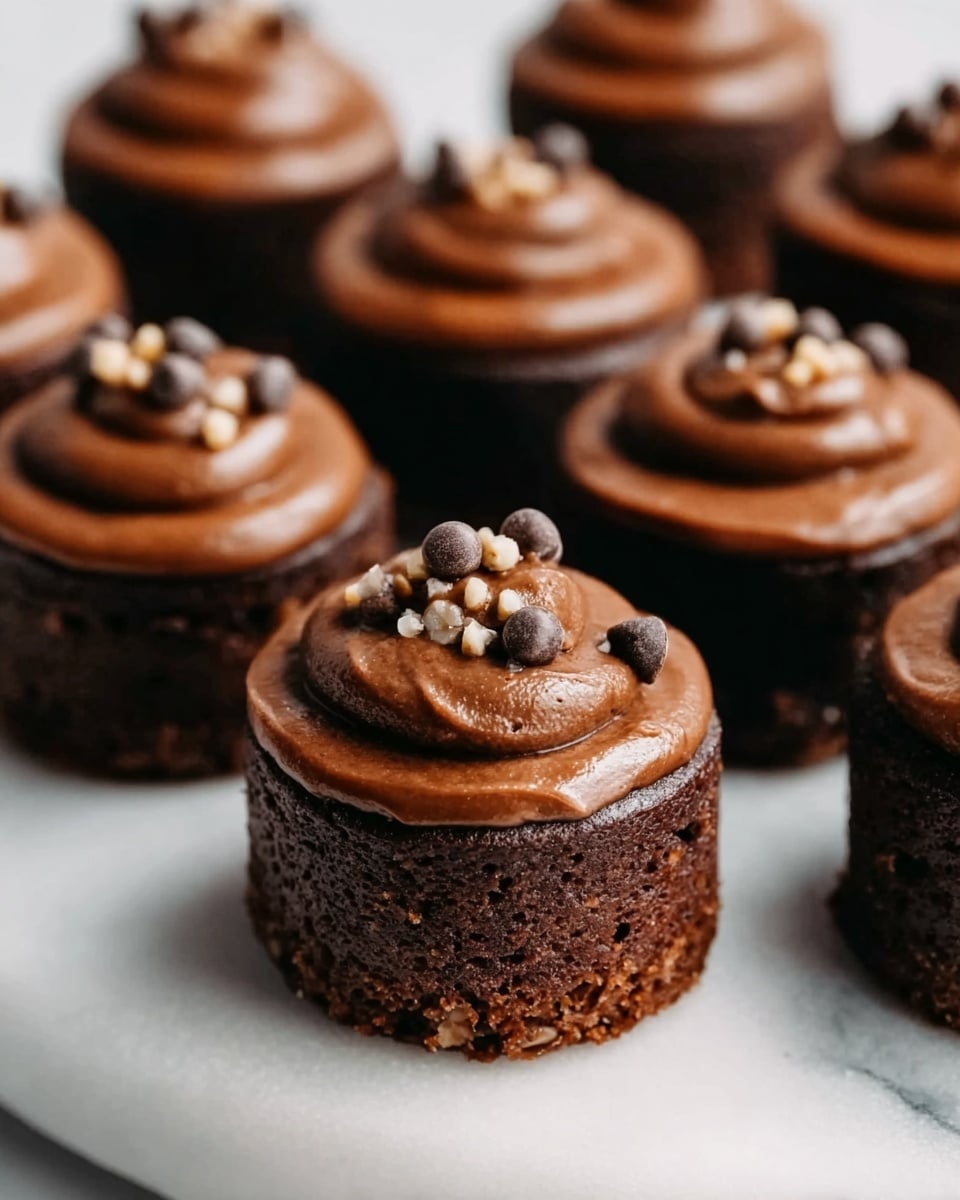 The image shows small, round chocolate cakes placed on a white marble surface. Each cake has two layers: a dark brown, dense base layer with a moist and slightly rough texture, topped with a smooth, creamy chocolate frosting. On top of the frosting, there are small pieces of chocolate and nuts, adding texture and detail. The cakes are evenly spaced, with one in the front clearly in focus and the others softly blurred in the background. Photo taken with an iphone --ar 4:5 --v 7