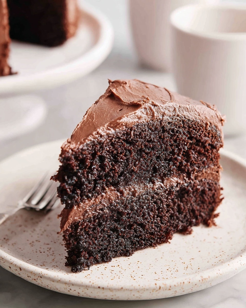 A slice of two-layer dark chocolate cake with rich, thick chocolate frosting spread on the top and between the layers, showing a moist and slightly crumbly texture. The cake slice sits on a white plate with small black specks, placed on a white marbled surface. The frosting is smooth but slightly uneven on the edges, emphasizing a homemade feel. In the blurred background, there are white cups adding soft contrast. photo taken with an iphone --ar 4:5 --v 7