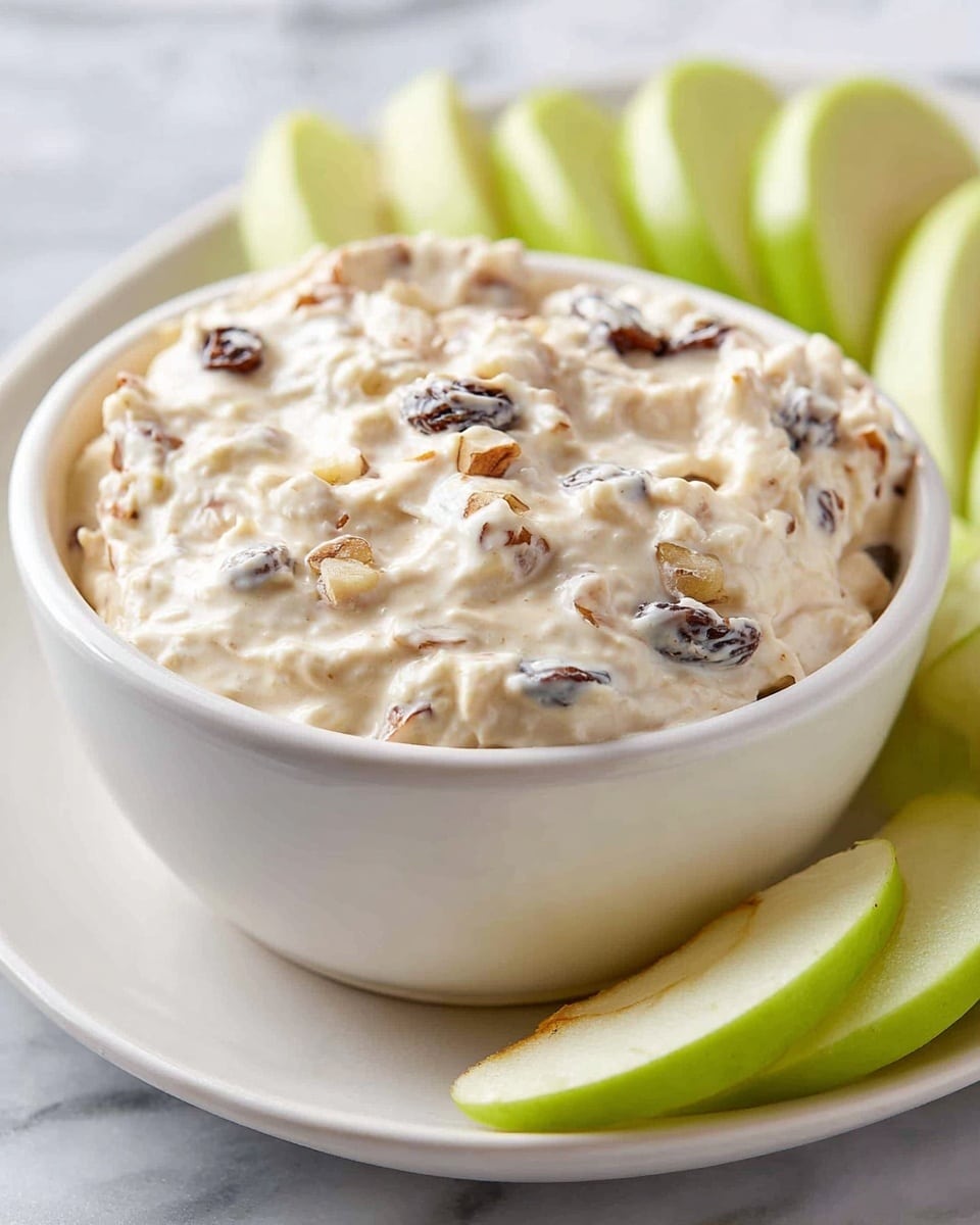 A close-up view of a white bowl filled with creamy, thick dip mixed with small pieces of dark raisins and light brown nuts, showing a textured surface with soft peaks and swirls. The bowl sits on a matching white plate, surrounded by fresh green apple slices arranged partially out of focus in the background on a white marbled surface. The overall colors are light cream with dark and tan bits, contrasted by the bright green of the apple slices. photo taken with an iphone --ar 4:5 --v 7