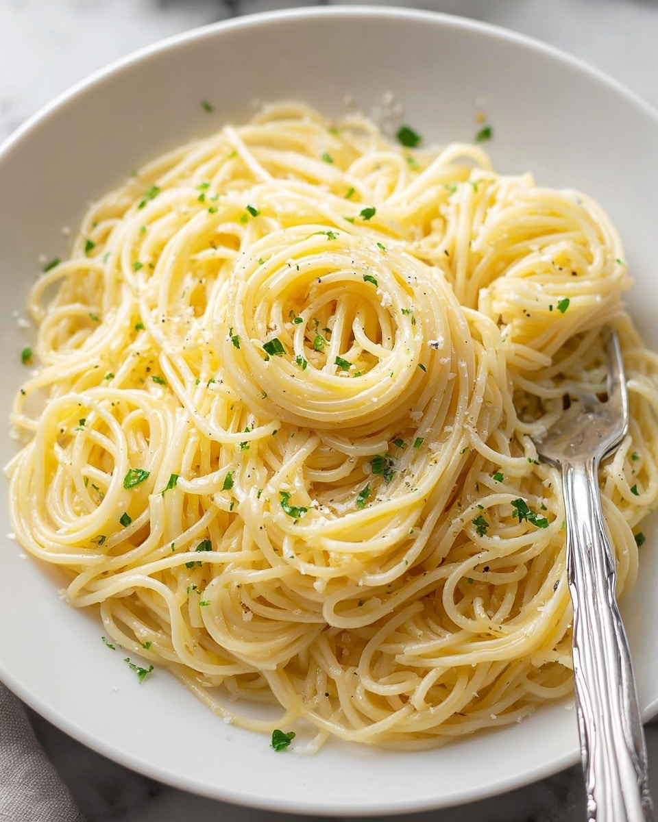 A white plate with a small pile of spaghetti noodles in the center, each strand pale yellow and slightly glossy, showing light oil or butter coating. The noodles are twirled loosely, with some overlapping and curling at the edges. Small green bits of parsley are scattered evenly on top, mixed in with tiny white shavings of cheese and small black pepper specks. A silver fork rests inside the plate on the right side next to the pasta. The background and surface are a white marbled texture. photo taken with an iphone --ar 4:5 --v 7