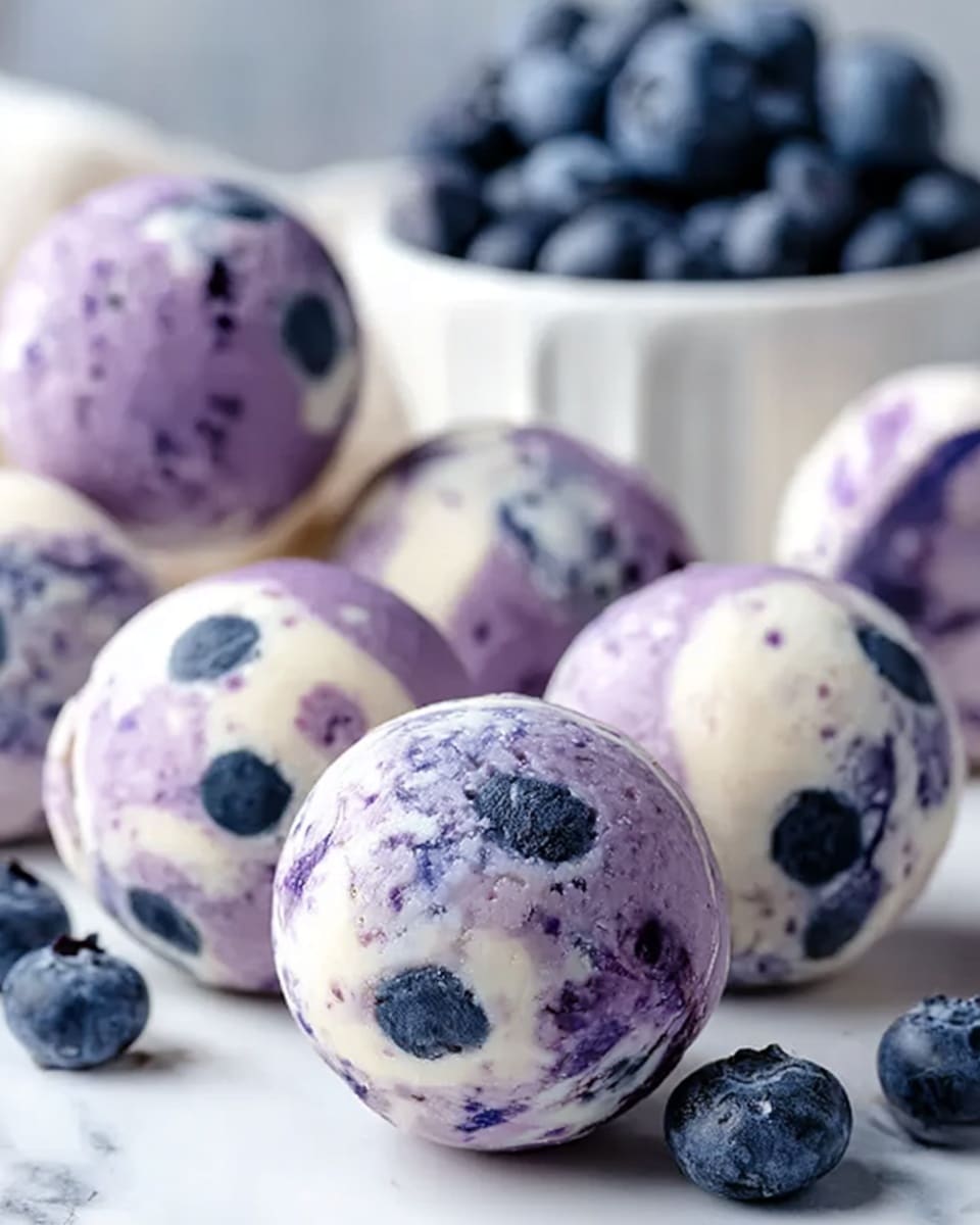 The image shows several round dessert balls with a smooth, creamy texture. Each ball has a white base with shades of purple mixed inside, creating a marbled effect. There are whole blueberries embedded within the balls, adding dark blue spots that contrast with the lighter creamy parts. The balls are arranged on a white marbled surface, with a white bowl filled with more blueberries blurred in the background. Photo taken with an iphone --ar 4:5 --v 7