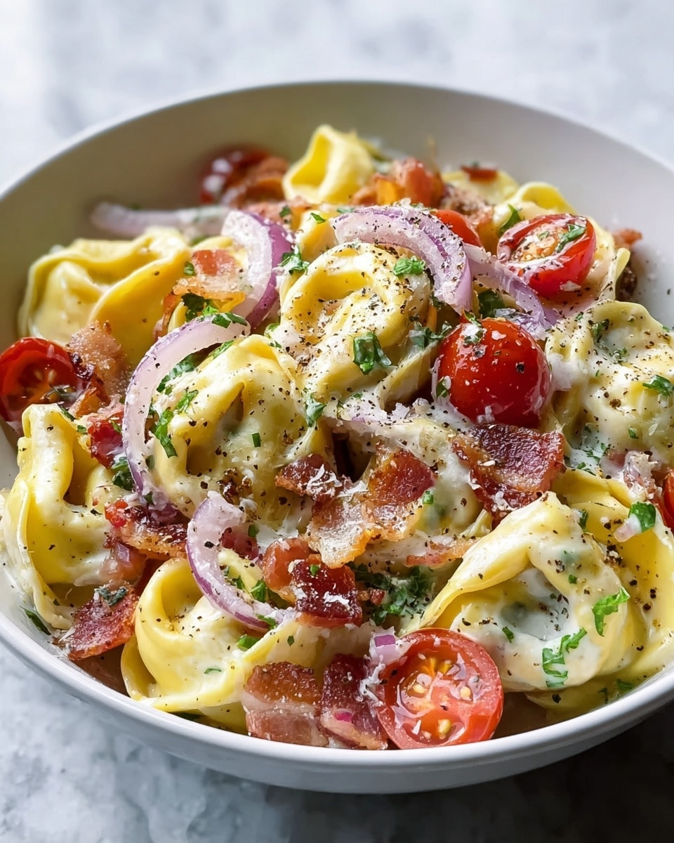 A close-up view of a white bowl filled with creamy tortellini pasta layered with folded pale yellow pasta filled with cheese, coated in a rich white sauce. Mixed in are halved bright red cherry tomatoes, thin rings of light purple onion, and crispy brown bits of bacon scattered on top. The dish is sprinkled with small green parsley pieces and a light dusting of black pepper. The bowl sits on a white marbled surface with part of a gray cloth visible beneath it. Photo taken with an iphone --ar 4:5 --v 7