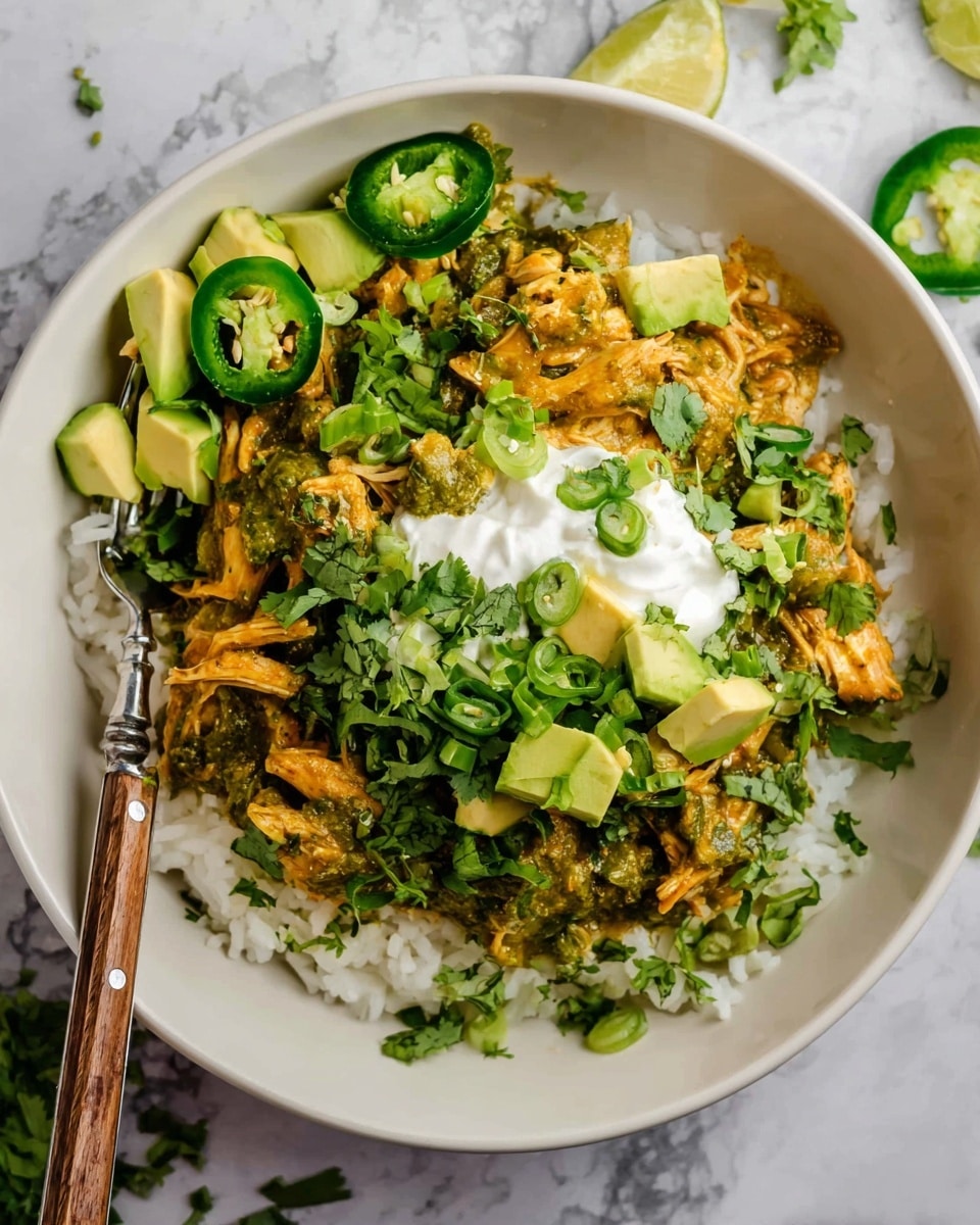 A white bowl filled with a base layer of white rice, topped with shredded chicken cooked in a green herb sauce, giving it a slightly orange-green color and moist texture. On top of the chicken, there are small cubes of bright green avocado and fresh green cilantro leaves scattered around. A dollop of white sour cream sits in the center with a few chopped green onions sprinkled on it. Thin slices of green jalapeño peppers are placed on the side of the bowl. A silver fork with a wooden handle rests on the edge of the bowl on a white marbled surface. Photo taken with an iphone --ar 4:5 --v 7