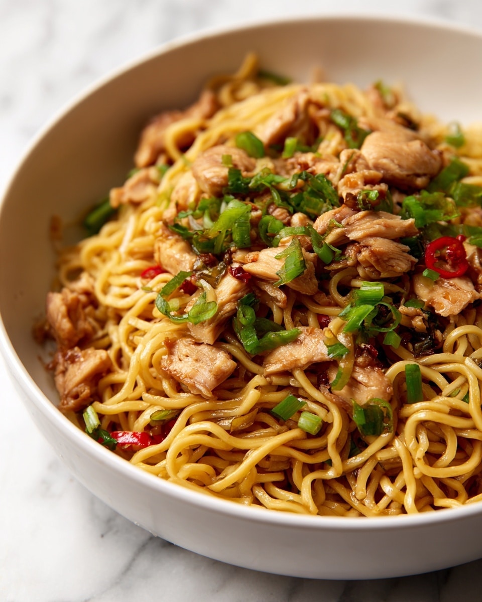 The dish shows a close-up of cooked noodles mixed with several small chunks of light brown cooked chicken, scattered green chopped scallions, and small pieces of red bell pepper. The noodles are golden yellow, slightly glossy with sauce, forming the base layer, with chicken pieces evenly spread on top. The scallions and red pepper bits sit on the surface, adding bright green and red colors and fresh texture. The food is served in a white bowl set against a white marbled surface. The image focuses tightly on the food, showing the shiny, moist texture of the ingredients. Photo taken with an iphone --ar 4:5 --v 7