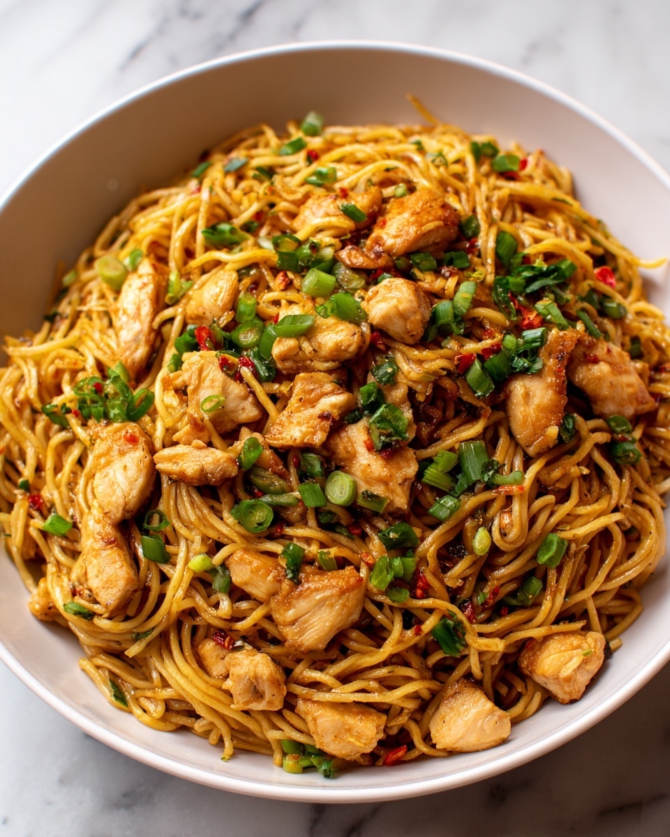 A black bowl is filled with a large serving of cooked noodles mixed with small pieces of light brown chicken. The noodles are golden-yellow and slightly curly, with finely chopped green onions and small red pepper pieces scattered throughout. On top, there is a sprinkle of chopped fresh green herbs, adding a pop of color and freshness. The bowl sits on a white marbled surface with a blurred background showing other dishes. photo taken with an iphone --ar 4:5 --v 7