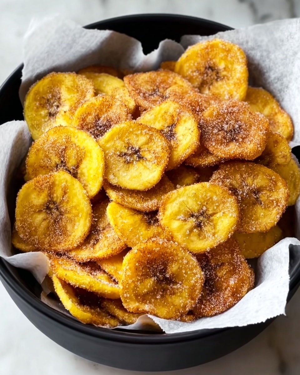 A basket lined with white paper holds a pile of golden-yellow plantain chips dusted with fine white sugar and cinnamon powder. The chips are thin, round, and slightly browned in spots, showing a crispy texture. The basket has a black edge and is placed on a white marbled surface. photo taken with an iphone --ar 4:5 --v 7