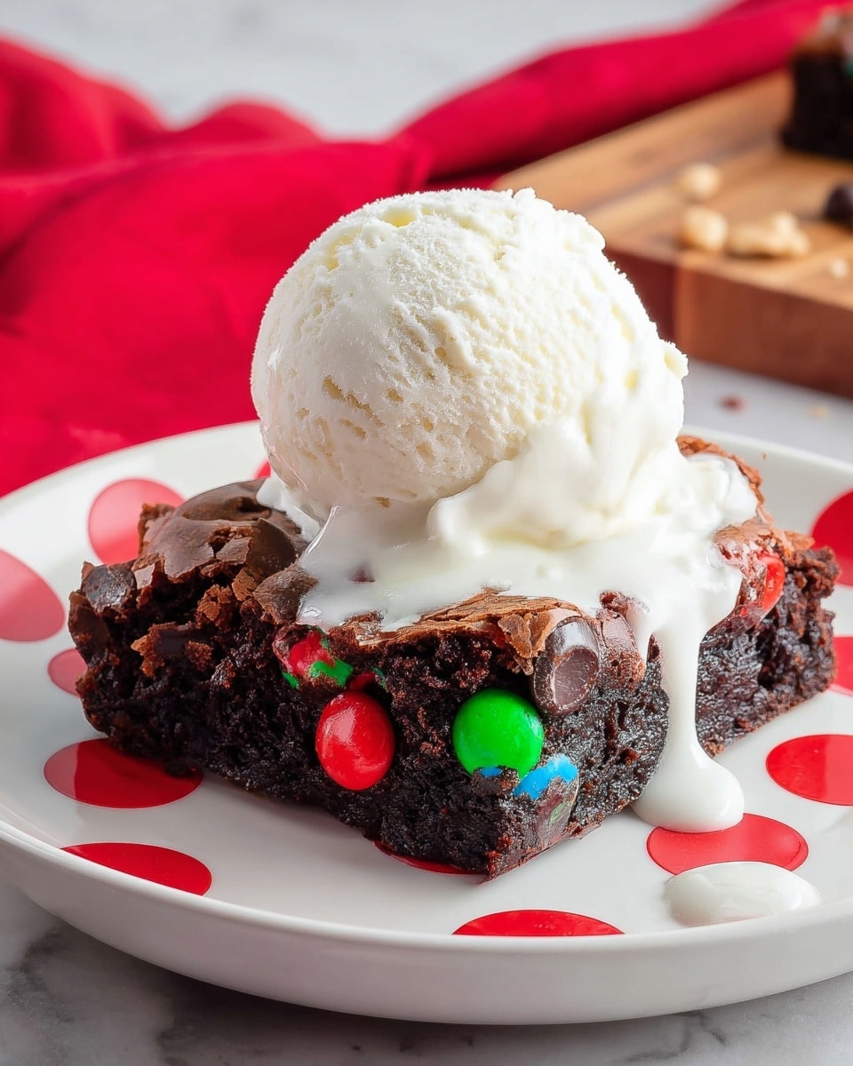 A single square brownie piece with a dark, rich, and moist texture forms the base, embedded with colorful red, green, and blue candy-coated chocolates and dark chocolate chips on top. A large scoop of white vanilla ice cream sits on the brownie, slightly melting and dripping down the sides. The dessert is placed on a white plate with bright red polka dots, sitting on a white marbled surface with a soft red cloth and a wooden board blurred in the background. Photo taken with an iphone --ar 4:5 --v 7