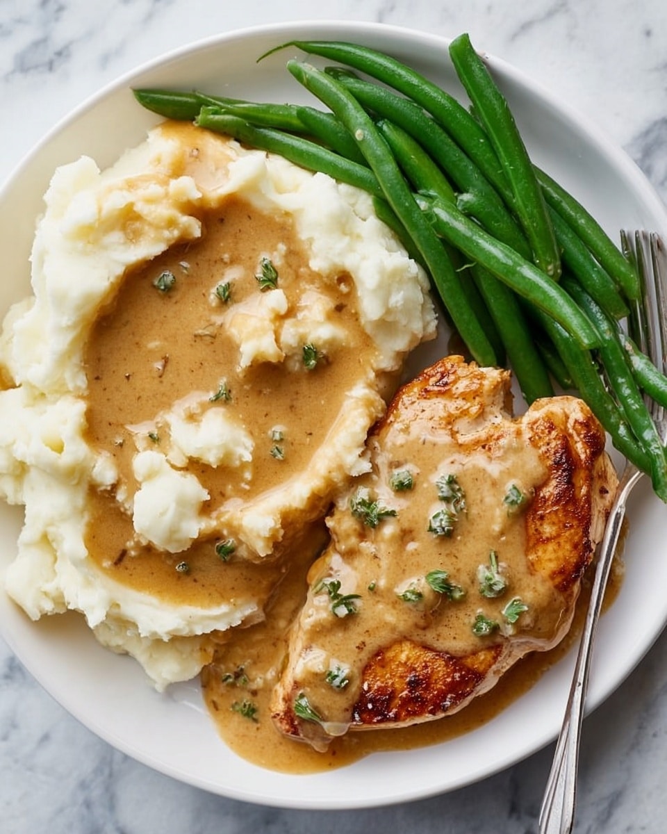 A white round plate holds a meal with three main parts. On the left, there is a golden brown grilled chicken breast covered with a creamy, light brown gravy that has small green herb bits on top. Next to it on the right is a thick mound of smooth, white mashed potatoes with black pepper sprinkled on top, partly covered with the same light brown gravy; a silver spoon is digging into the potatoes. At the top right of the plate, there is a neat pile of bright green cooked green beans, arranged side by side. The whole plate sits on a white marbled surface. Photo taken with an iphone --ar 4:5 --v 7