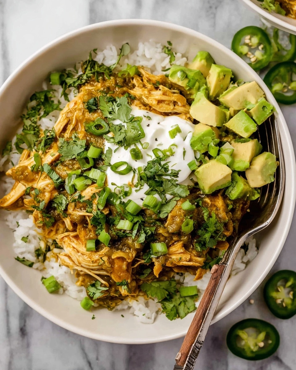 A white bowl with three main layers is shown: the bottom layer is white rice, the middle layer is orange-brown shredded chicken mixed with green herbs and sauce, and the top layer includes a dollop of white sour cream, bright green avocado cubes, sliced green jalapeños, and fresh green cilantro leaves scattered around. A silver fork with a wooden handle rests inside the bowl on the right side. The bowl is placed on a white marbled surface with scattered green herbs nearby. photo taken with an iphone --ar 4:5 --v 7