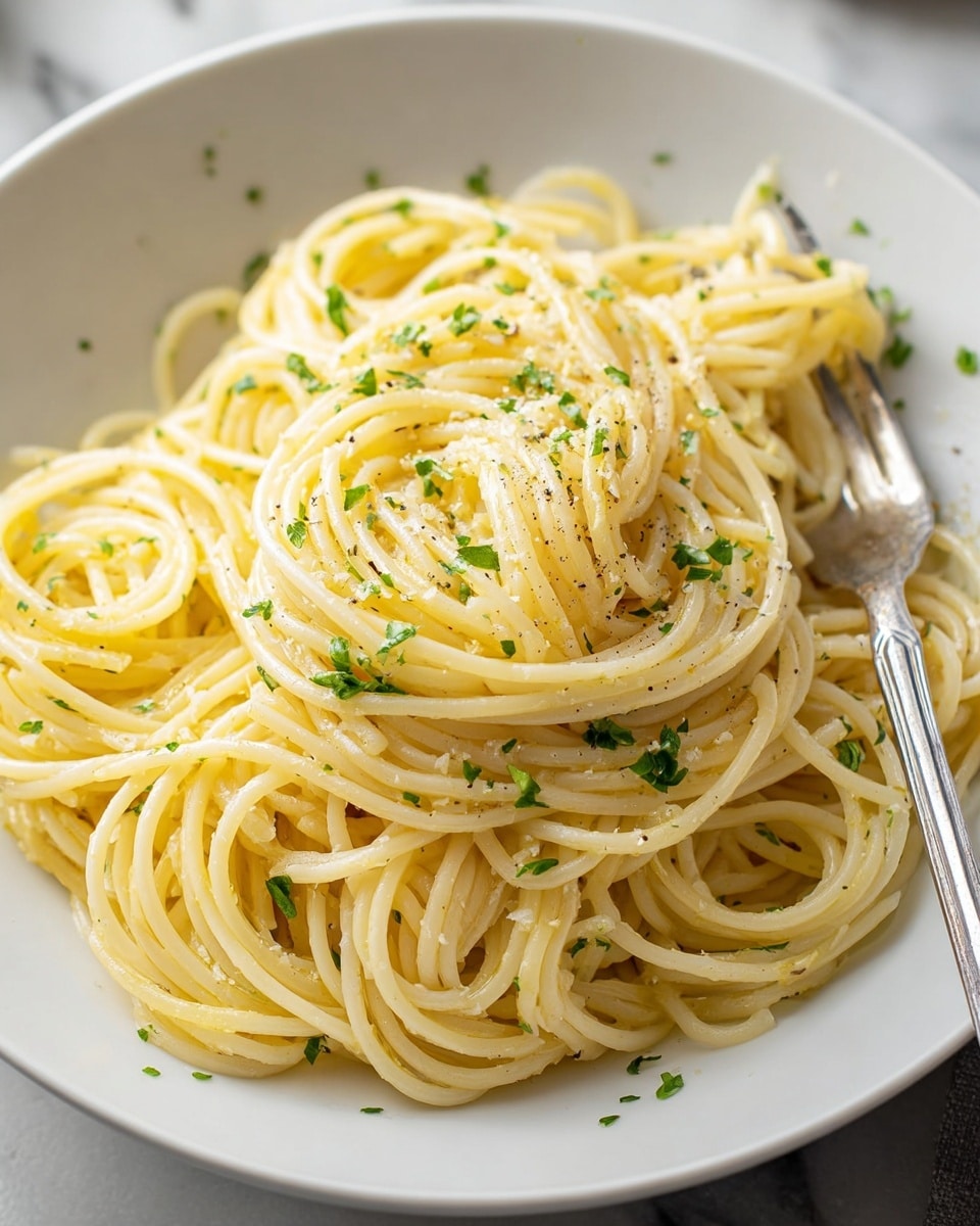 A white bowl filled with a neat mound of plain spaghetti noodles tossed lightly in oil or butter, giving them a soft, slightly shiny yellow color. The noodles are thin and smooth, arranged in loose coils creating depth and texture. Small bits of chopped green parsley and black pepper are sprinkled evenly over the top, adding subtle green and black specks. A fork with a shiny silver handle rests inside the bowl on the right side, partially visible. The background shows a blurred white marbled surface. Photo taken with an iphone --ar 4:5 --v 7