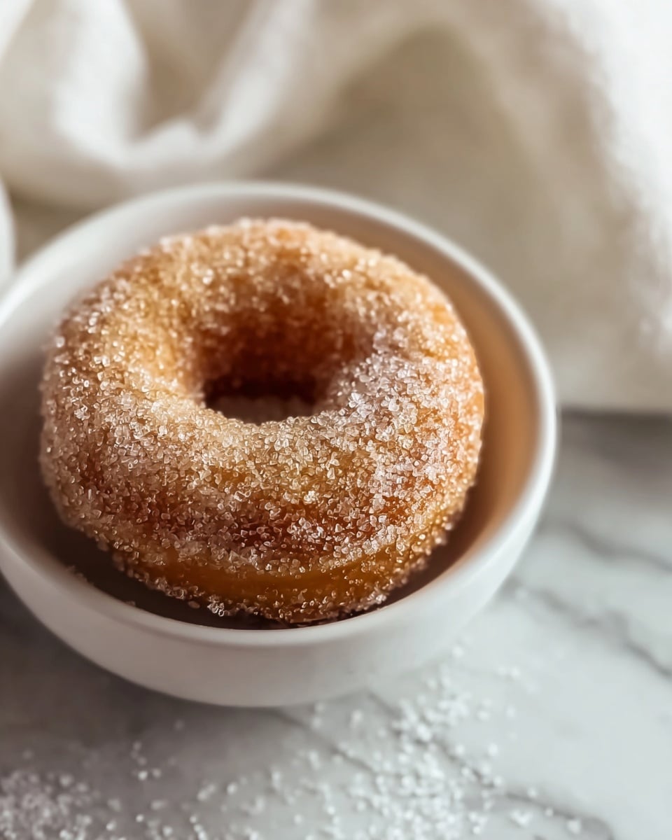 A close-up view of a single round cinnamon sugar donut with a hole in the center, sitting inside a white bowl. The donut is covered evenly with large sugar crystals, giving it a sparkling texture in shades of light brown and golden. The bowl rests on a white marbled surface scattered with more sugar crystals. The background shows a soft, white textured cloth, creating a cozy and inviting scene. Photo taken with an iphone --ar 4:5 --v 7