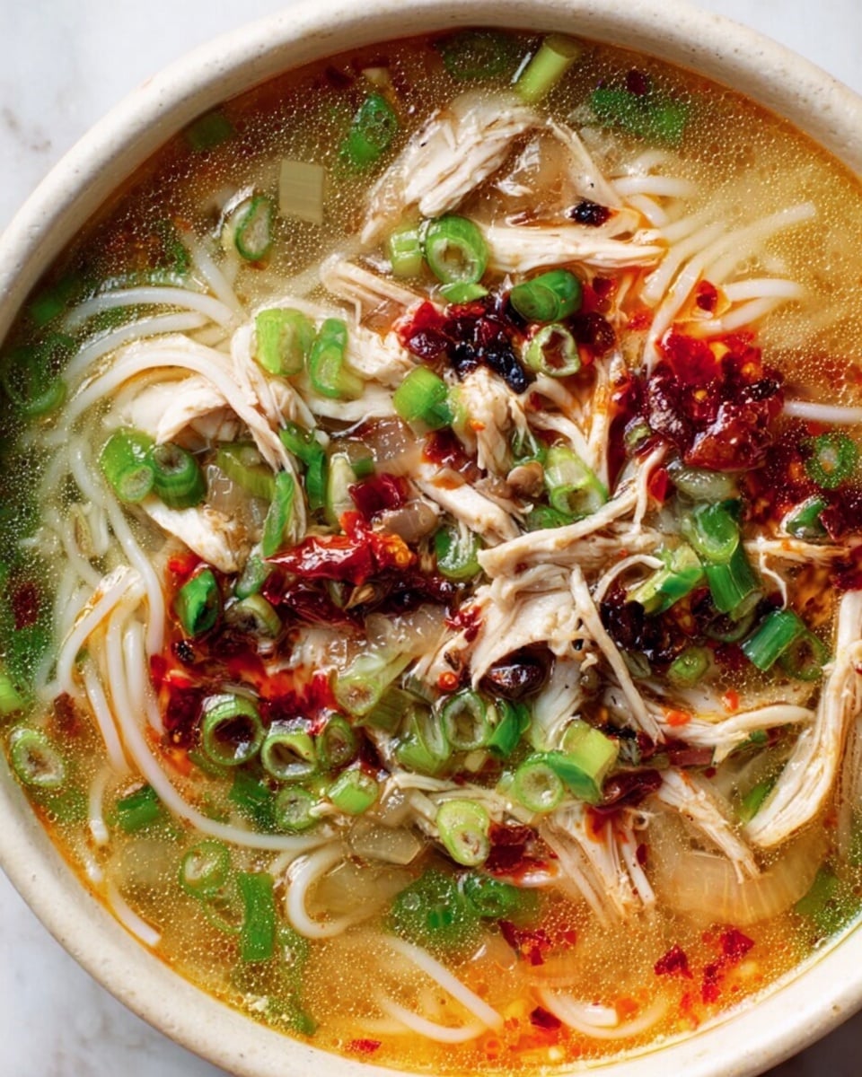 A close-up of a bowl of noodle soup with multiple layers visible: thin white noodles sit at the bottom, topped by shredded pieces of light brown chicken. Bright green sliced scallions are scattered across the top, along with bits of red chili oil that add splashy red and dark spots. The broth is a light yellowish color with oily patches and small pieces of cooked vegetables like translucent onions. The bowl is white and placed on a white marbled surface. Photo taken with an iphone --ar 4:5 --v 7