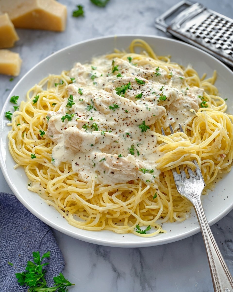 A white plate is filled with thin yellow spaghetti noodles as the bottom layer, loosely coiled around the edges. On top, slices of white creamy chicken coated in a thick off-white sauce with a smooth and slightly lumpy texture spread evenly across the middle. Small pieces of green parsley and specks of black pepper scatter over the top, adding a fresh touch. A silver fork is inserted into the noodles on the right side of the plate, partially lifting some strands. The dish is set on a white marbled surface, with a cheese grater and a few pieces of cheese visible in the background. photo taken with an iphone --ar 4:5 --v 7