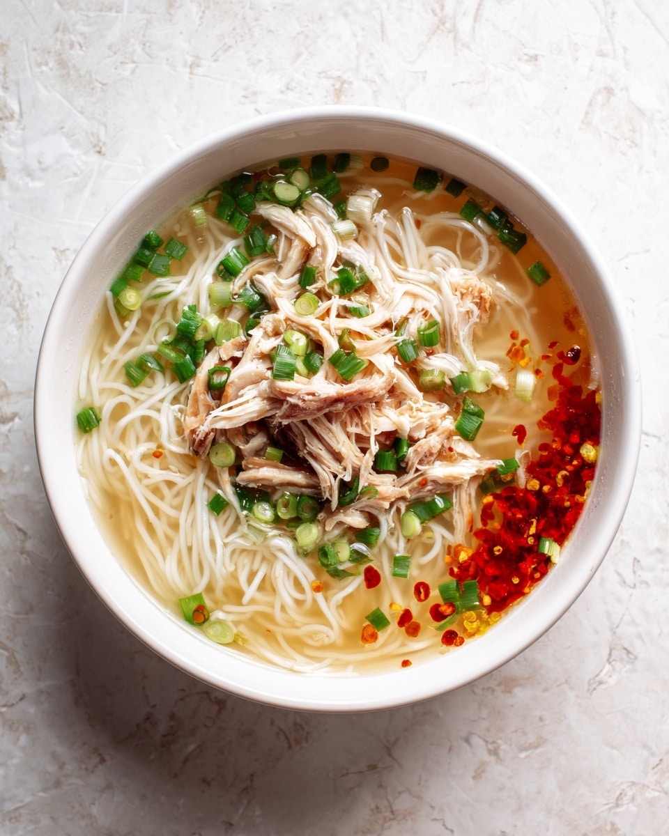The image shows a bowl of noodle soup with clear broth. Inside the bowl, there are thin white noodles filling most of the space. On top of the noodles, there are small pieces of shredded chicken placed in the middle, surrounded by sliced green onions scattered evenly. There are drops of red chili oil floating on the broth, giving bright red spots near the top right area of the bowl. The bowl is white and placed on a white marbled textured surface. photo taken with an iphone --ar 4:5 --v 7