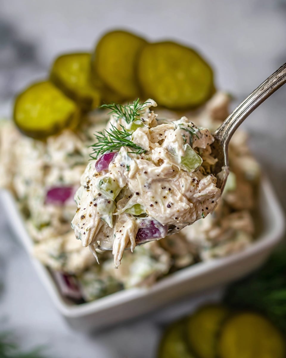 A close-up view of a spoonful of creamy chicken salad showing three main layers: shredded white chicken pieces mixed with creamy white dressing speckled with black pepper, small green herb sprigs, and chunks of purple-red onion. The spoon holds a generous scoop with visible fresh dill on top and bits of celery or pickle mixed in, creating a textured, chunky look. In the background, a square white bowl is filled with more creamy chicken salad and topped with several slices of green pickles arranged near the edge. The scene is set on a white marbled texture. Photo taken with an iphone --ar 4:5 --v 7