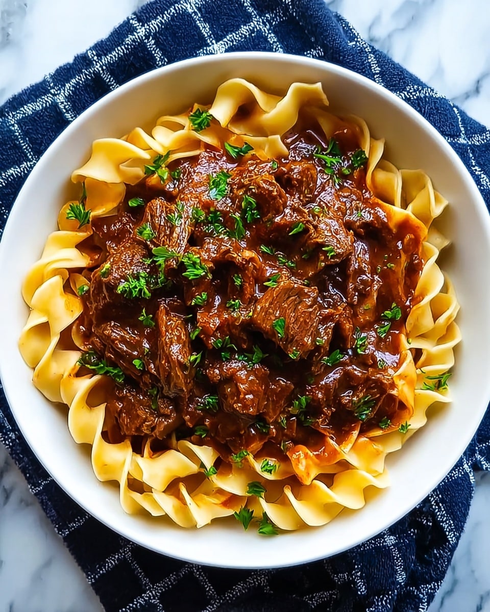 The image shows a white bowl filled with a rich, dark brown beef and noodle dish. The base layer consists of wide, flat yellow noodles coated lightly in sauce, winding through the dish. On top of the noodles, there is a generous amount of tender, chunky pieces of beef, covered in a thick, glossy brown sauce. Scattered across the beef and noodles are small green slices of herbs which add a fresh touch. The bowl sits on a white marbled surface with a hint of a blue and white cloth underneath. The focus is close, capturing the textures and shine of the sauce and the softness of the meat and noodles. photo taken with an iphone --ar 4:5 --v 7