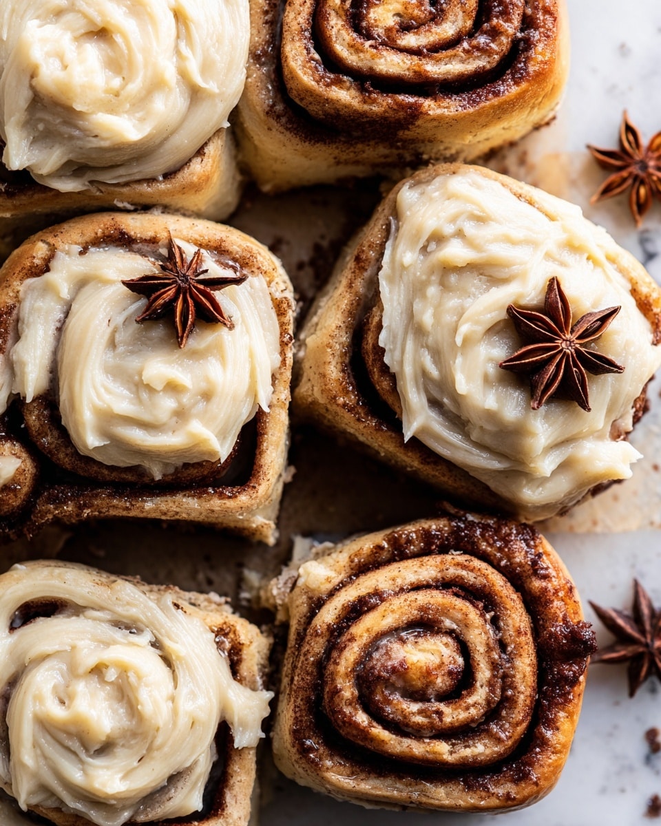 A close-up top view of six cinnamon rolls arranged in a cluster on a white marbled surface. Each roll has a visible spiral shape with a dark brown cinnamon layer forming distinct swirls inside the lighter beige dough. The rolls are generously topped with a creamy, rich beige frosting that is thick and swirled unevenly, covering parts of the cinnamon spirals. Near the center, there is a whole star anise placed on one of the rolls, adding a decorative touch. The texture of the rolls looks soft and slightly crumbly, with some icing dripping down the sides. The scene is warm and inviting, showcasing the cinnamon rolls up close. photo taken with an iphone --ar 4:5 --v 7