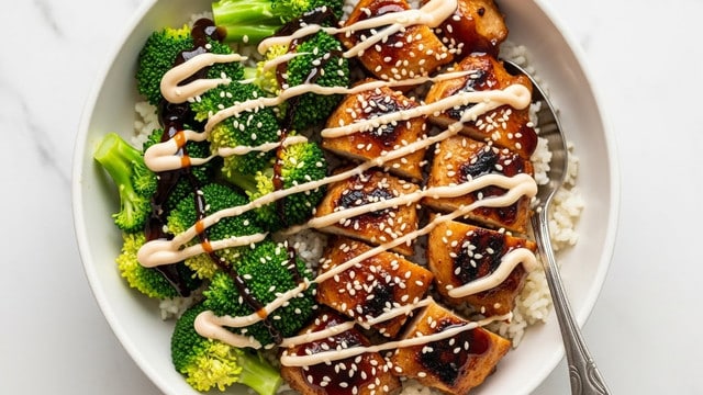 The image shows a white bowl filled with three main layers: at the bottom is a thick layer of white rice with a soft, fluffy texture; on top of the rice are golden-brown, grilled chicken pieces that look juicy and slightly charred, covering about half of the bowl; next to the chicken, bright green broccoli florets are placed, looking fresh and slightly steamed; the whole dish is drizzled with a creamy light beige sauce scattered unevenly over the chicken and broccoli, with some sesame seeds sprinkled on top, adding texture. A silver fork rests on the right side, partially inserted into the rice and chicken, and the setting is on a white marbled texture. photo taken with an iphone --ar 4:5 --v 7