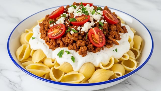 A white bowl with a blue rim holds a three-layer dish on a white marbled texture. The bottom layer is large, shell-shaped pasta, light yellow with a smooth texture. The middle layer is a creamy white sauce spread evenly on top of the pasta. The top layer is a mix of browned ground meat with onions in a rich reddish-brown sauce. Bright red cherry tomato halves and small bits of white cheese are scattered over the meat, along with chopped green herbs sprinkled on the entire dish. photo taken with an iphone --ar 4:5 --v 7