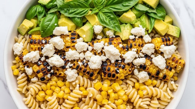 The image shows a close-up of a white bowl filled with a pasta salad. The dish has three main layers: the base layer is light beige spiral pasta with a slightly firm texture, mixed with small bright yellow corn kernels; the middle layer has charred corn pieces with dark brown grill marks and creamy white crumbles of cheese spread evenly; the top layer includes fresh green basil leaves and light green avocado chunks. The ingredients are mixed but still visible in layers and placed on a white marbled texture background. Photo taken with an iphone --ar 4:5 --v 7