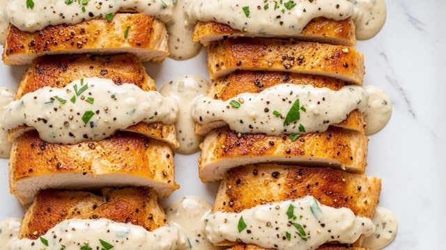 The image shows several pieces of golden-brown cooked chicken breast, cut into thick slices, lying close together. Each piece is covered in a creamy, white sauce that has small black pepper and green herb specks visible on the surface. The sauce looks smooth and slightly thick, pooling around the chicken. The chicken pieces have a slightly crispy texture on the edges, and the sauce’s creamy texture contrasts with the seared chicken. The dish is in a white container or pan, all set on a white marbled texture background. photo taken with an iphone --ar 4:5 --v 7