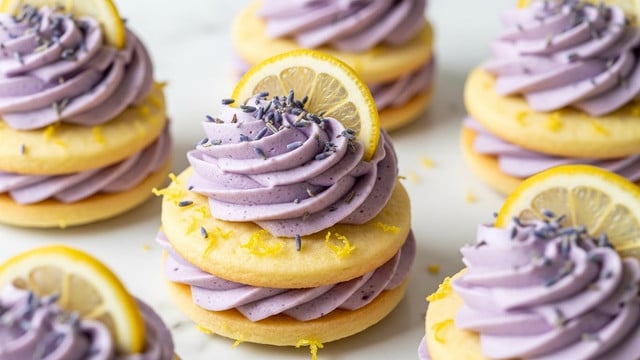 The image shows lemon lavender sandwich cookies placed on a white marbled surface. Each cookie is made of two pale yellow round cookie layers with a smooth texture, sandwiching a thick layer of light purple lavender cream in between. On top of each sandwich, there is a generous swirl of the same lavender cream, decorated with a thin, semi-transparent lemon slice and small lavender buds scattered over the cream. The lemon cookie surfaces are sprinkled lightly with lemon zest, adding a touch of texture and bright color. Photo taken with an iphone --ar 4:5 --v 7