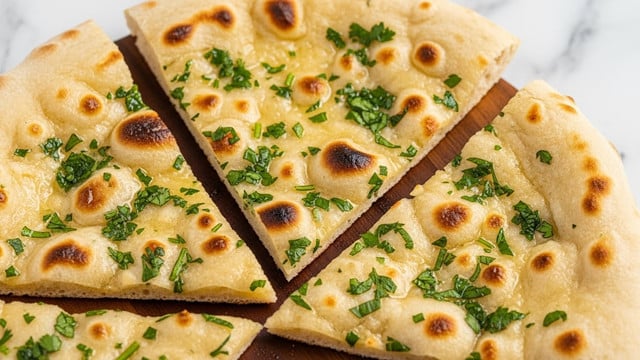 A close-up image showing three slices of flatbread named naan, each slice with a thin crust colored light golden with small darker toasted spots evenly spread across. The flatbread is decorated with scattered green herbs finely chopped on top. The texture looks soft, fluffy, and lightly oily. The slices are placed on a wooden surface with a white marbled texture in the background. Photo taken with an iphone --ar 4:5 --v 7