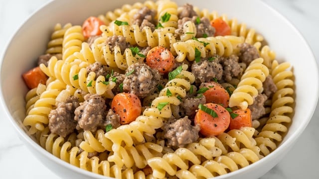 A close-up view of a black bowl filled with creamy baked pasta showing about three layers: the bottom layer is a smooth, white cream sauce; the middle layer consists of yellow spiral pasta mixed with small browned bits of sausage; the top layer is melted light golden cheese with toasted brown spots and scattered green herb leaves. The bowl rests on a dark cloth set on a white marbled surface. photo taken with an iphone --ar 4:5 --v 7