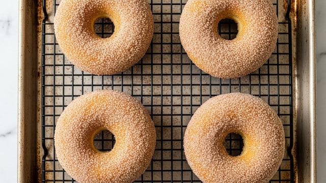 Four donuts are sitting on a black wire cooling rack, which is placed on a metal baking sheet with a slightly rusty look. Each donut is fully covered with a light brown sugar and cinnamon coating that gives a grainy texture. The donuts have a soft, golden-brown baked surface with a classic ring shape and a smooth yet slightly rough texture from the sugar. The background below the baking sheet is a white marbled texture. photo taken with an iphone --ar 4:5 --v 7