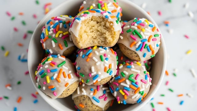 A white bowl filled with round, cake-like balls covered in white icing and brightly colored rainbow sprinkles, each ball smooth with a slightly speckled texture. One ball is positioned on top, with a big bite taken out, revealing a soft, crumbly pale yellow inside. The white bowl sits on a white marbled surface scattered with small pieces of the same rainbow sprinkles, creating a playful scene. photo taken with an iphone --ar 4:5 --v 7